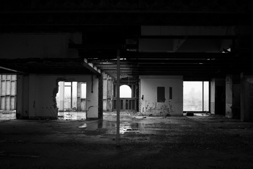 
Black and white photograph. 
The derelict building and the roof appear to open up and the rain has collected on the floor. You can see the reflection in the puddles. Further at the back of the open space, small windows leak in light from the outside. There’s one thin pillar holding the roof in the center of the frame and a wall that has been bashed.
