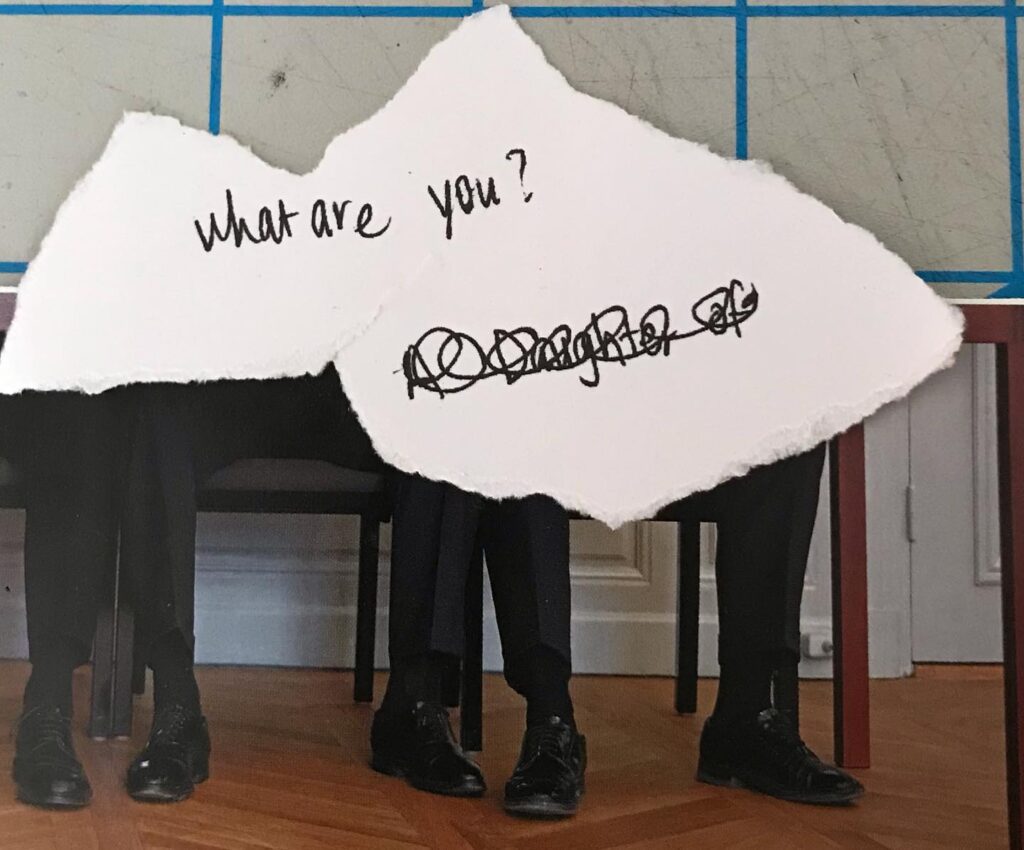 a surreal analog collage of 3 and a half people’s feet sitting on chairs with a torn white paper that says, “what are you?” With black scribbles crossing out, “a daughter of”underneath.
