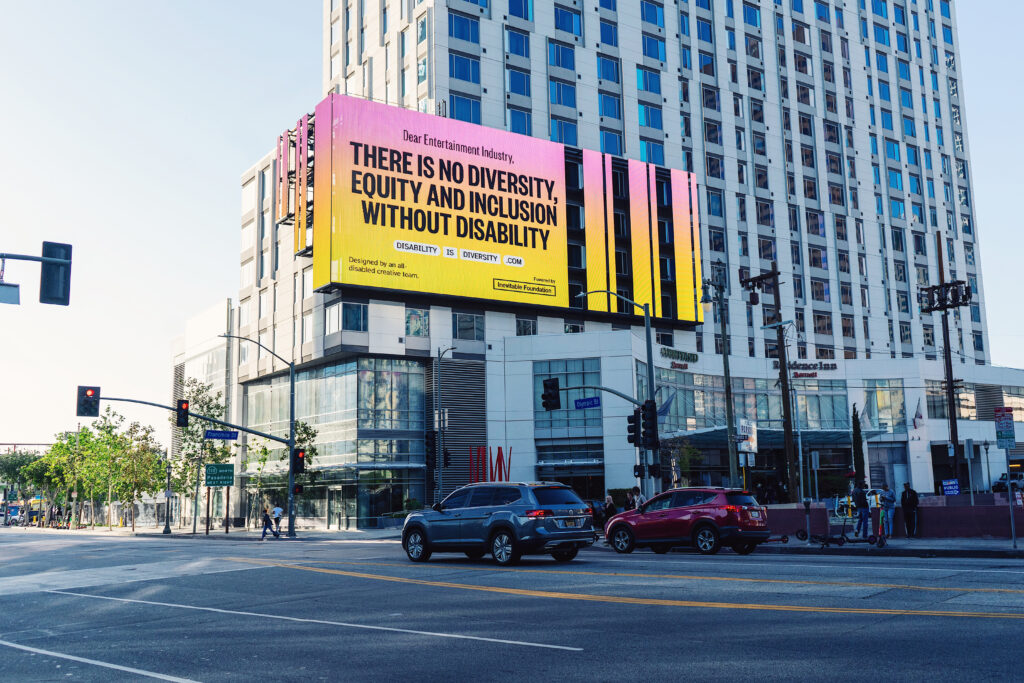 Photo of a billboard on a building that has bold text over a bright pink, orange and yellow gradient background that reads "There's no diversity, equity, and inclusion without disability." 