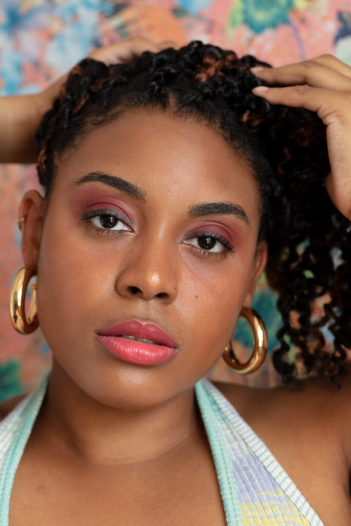Portrait photograph of a woman with a heavy hooped earrings. Her hair is in crochet braids and she is looking into the camera. She is wearing a light blue, lilac and yellow patterned vest top. She is wearing pink lipstick and maroon eyeshadow.